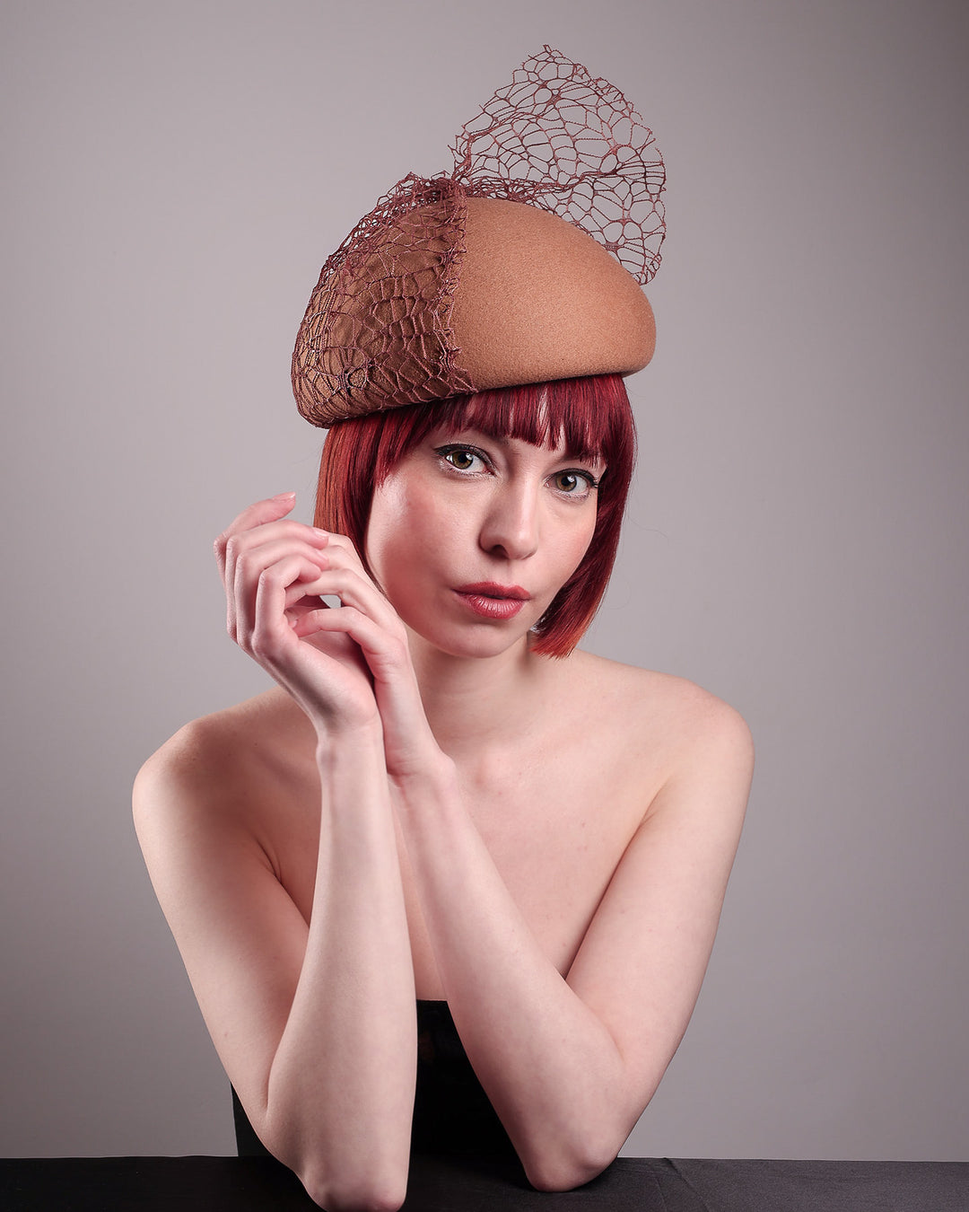 The model is showcasing the front right angle of the Lira Pillbox Beret Hat, which is a modern take on classic winter headwear. Crafted from warm brown wool felt, the softly rounded dome shape is elevated by an intricate mesh overlay on the right side, with a fan-like stand on the left side. Designed by Merve Bayindir. 