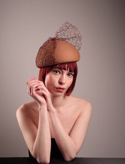 The model is showcasing the front right angle of the Lira Pillbox Beret Hat, which is a modern take on classic winter headwear. Crafted from warm brown wool felt, the softly rounded dome shape is elevated by an intricate mesh overlay on the right side, with a fan-like stand on the left side. Designed by Merve Bayindir. 
