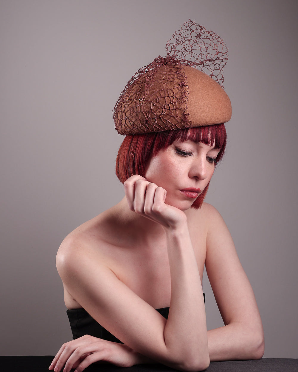 The model is showcasing the right front angle of the Lira Pillbox Beret Hat, which is a modern take on classic winter headwear. Crafted from warm brown wool felt, the softly rounded dome shape is elevated by an intricate mesh overlay on the right side, with a fan-like stand on the left side. Designed by Merve Bayindir. 
