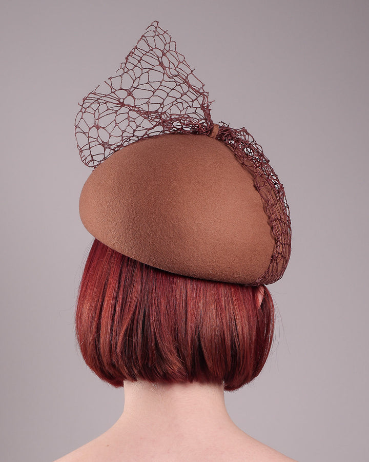 The model is showcasing the back of the Lira Pillbox Beret Hat, which is a modern take on classic winter headwear. Crafted from warm brown wool felt, the softly rounded dome shape is elevated by an intricate mesh overlay on the right side, with a fan-like stand on the left side. Designed by Merve Bayindir. 