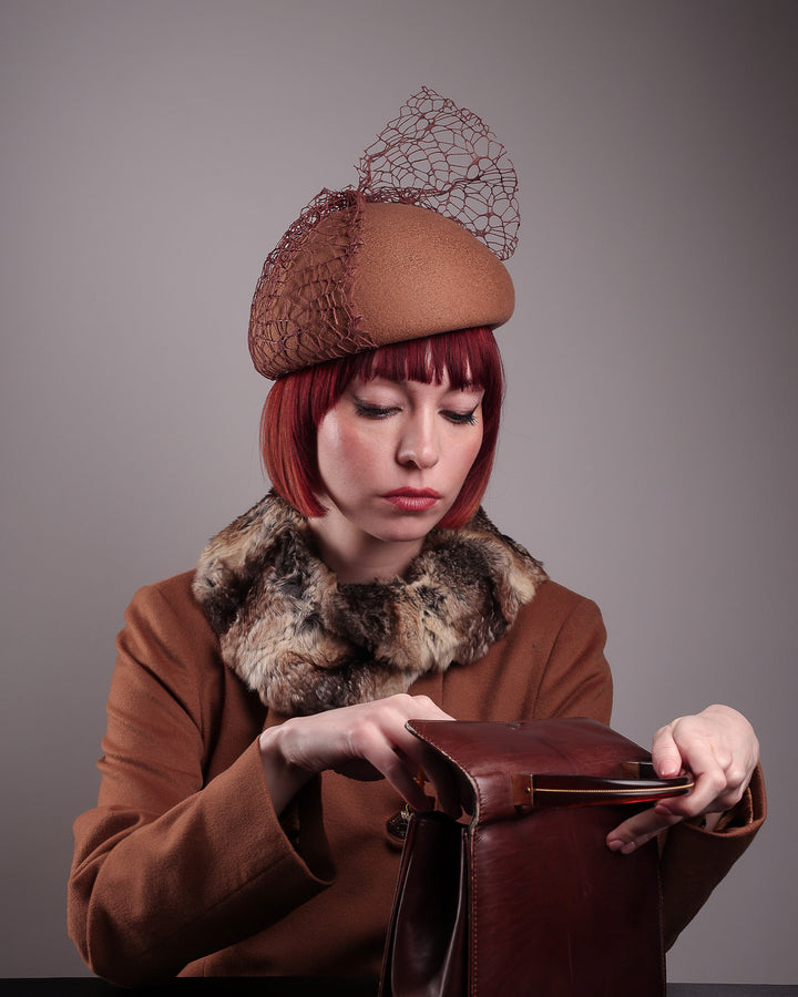 The model is showcasing the Lira Pillbox Beret Hat, which is a modern take on classic winter headwear. Crafted from warm brown wool felt, the softly rounded dome shape is elevated by an intricate mesh overlay on the right side, with a fan-like stand on the left side. This editoral picture is also showcases a brick coloured vintage coat and a vintage hand back to create a visual styling. Designed by Merve Bayindir. 