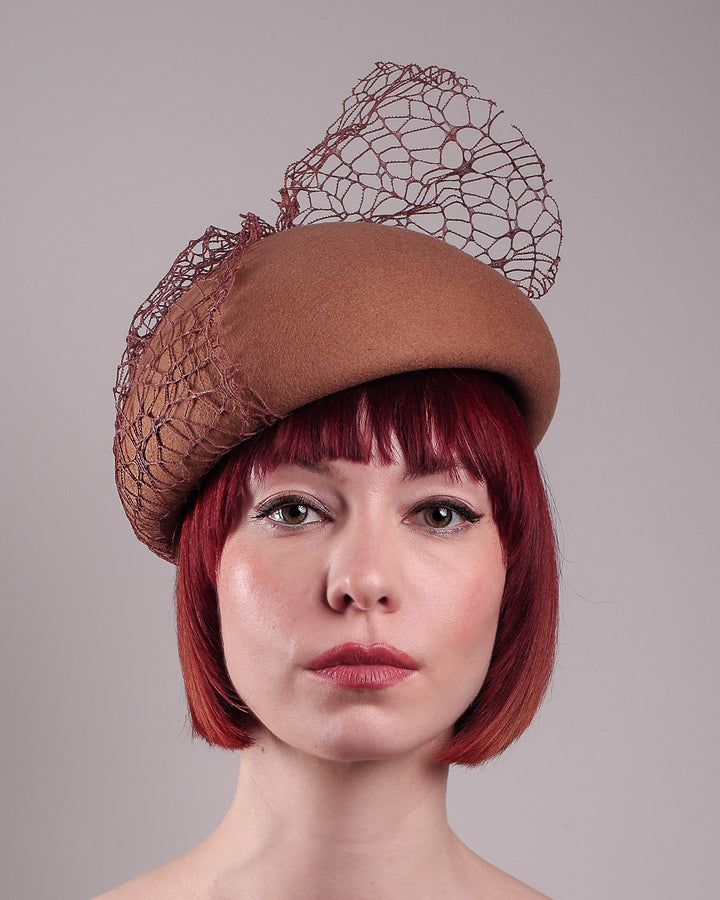 The model is showcasing the front close up of the Lira Pillbox Beret Hat, which is a modern take on classic winter headwear. Crafted from warm brown wool felt, the softly rounded dome shape is elevated by an intricate mesh overlay on the right side, with a fan-like stand on the left side. Designed by Merve Bayindir. 