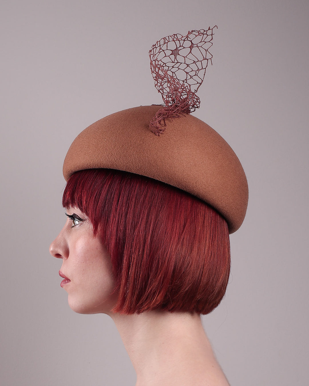 The model is showcasing the left side close up of the Lira Pillbox Beret Hat, which is a modern take on classic winter headwear. Crafted from warm brown wool felt, the softly rounded dome shape is elevated by an intricate mesh overlay on the right side, with a fan-like stand on the left side. Designed by Merve Bayindir. 