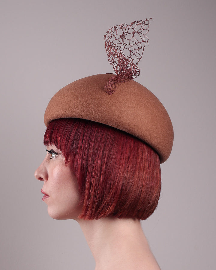 The model is showcasing the left side close up of the Lira Pillbox Beret Hat, which is a modern take on classic winter headwear. Crafted from warm brown wool felt, the softly rounded dome shape is elevated by an intricate mesh overlay on the right side, with a fan-like stand on the left side. Designed by Merve Bayindir. 