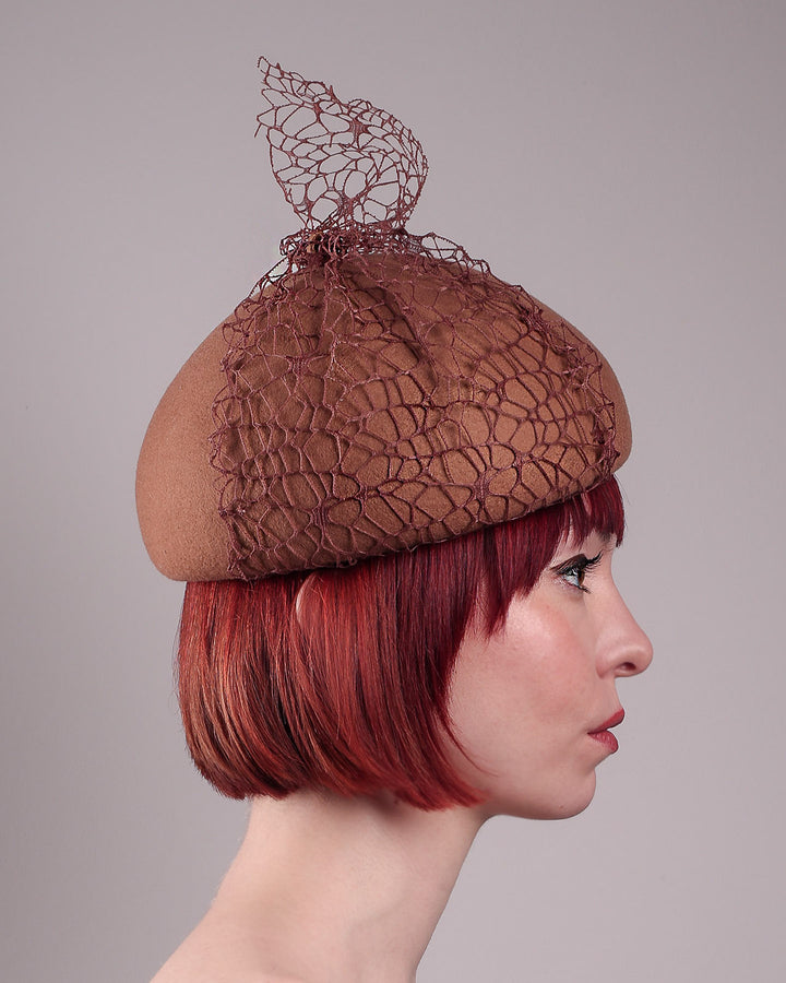 The model is showcasing the right side close up of the Lira Pillbox Beret Hat, which is a modern take on classic winter headwear. Crafted from warm brown wool felt, the softly rounded dome shape is elevated by an intricate mesh overlay on the right side, with a fan-like stand on the left side. Designed by Merve Bayindir. 