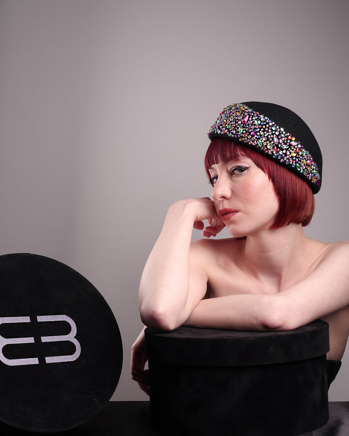 The editorial picture of the model with Merve Bayindir hat boxes while showcasing the left side of the Lumi Beret, crafted in classic black felt, and adorned with multi-coloured crystals that wrap across the front to the side of the brim. Designed by Merve Bayindir.