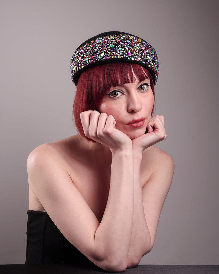 The model showcases the angled front of the Lumi Beret, crafted in classic black felt, and adorned with multi-coloured crystals that wrap across the front to the side of the brim. Designed by Merve Bayindir.