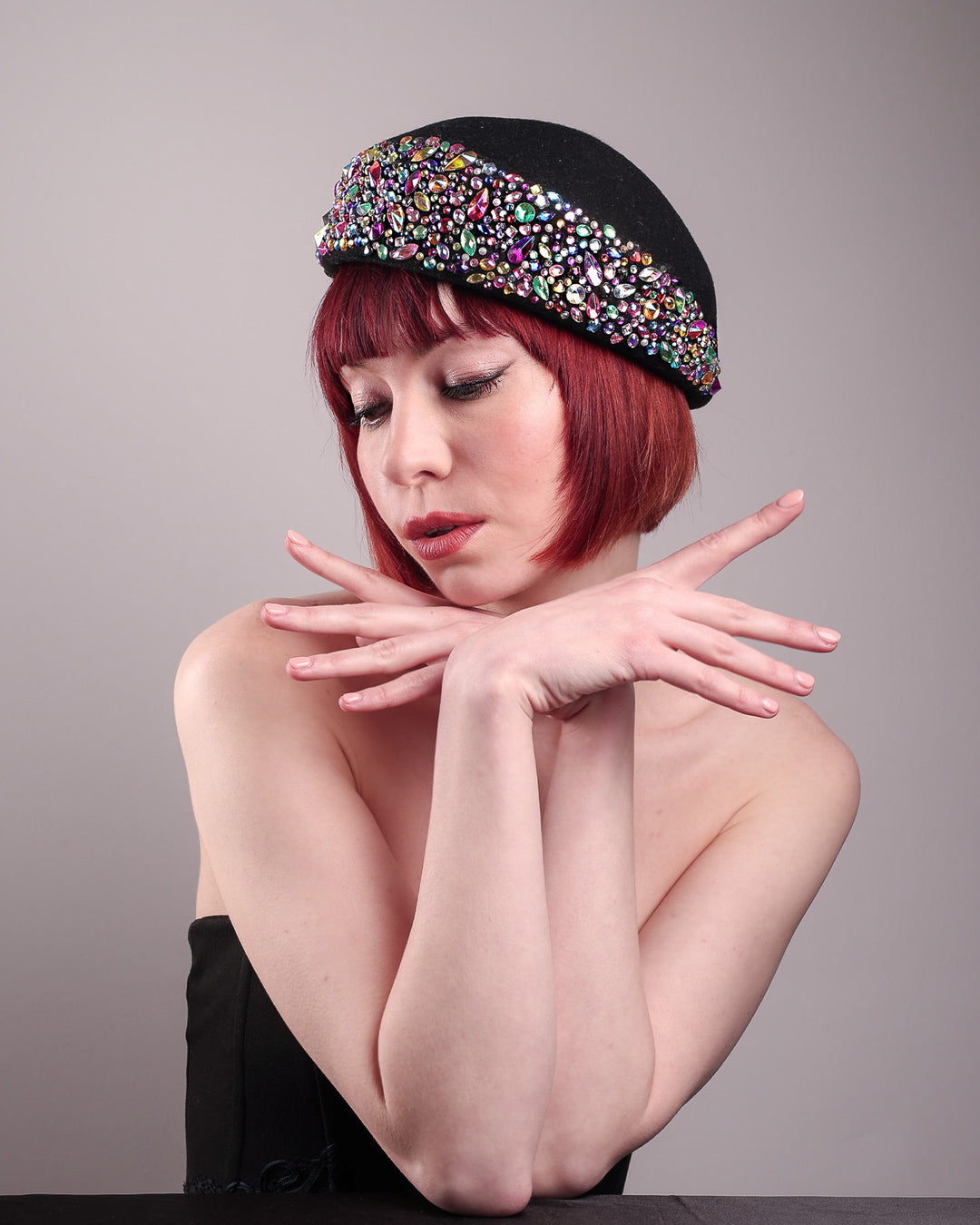 The model showcases the left side front of the Lumi Beret, crafted in classic black felt, and adorned with multi-coloured crystals that wrap across the front to the side of the brim. Designed by Merve Bayindir.