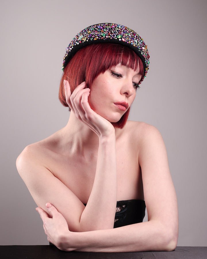 The model showcases the right side front of the Lumi Beret, crafted in classic black felt, and adorned with multi-coloured crystals that wrap across the front to the side of the brim. Designed by Merve Bayindir.