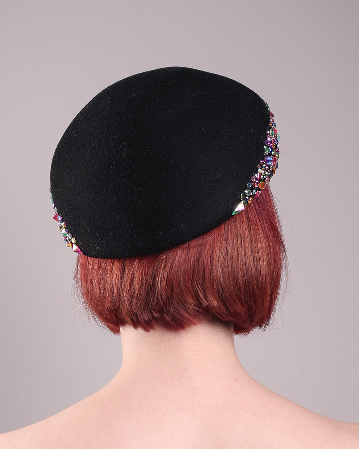 The model showcases the back of the Lumi Beret, crafted in classic black felt, and adorned with multi-coloured crystals that wrap across the front to the side of the brim. Designed by Merve Bayindir.