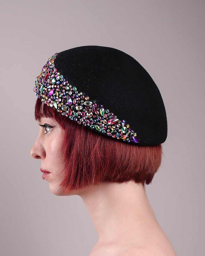 The model showcases the left side close up of the Lumi Beret, crafted in classic black felt, and adorned with multi-coloured crystals that wrap across the front to the side of the brim. Designed by Merve Bayindir.