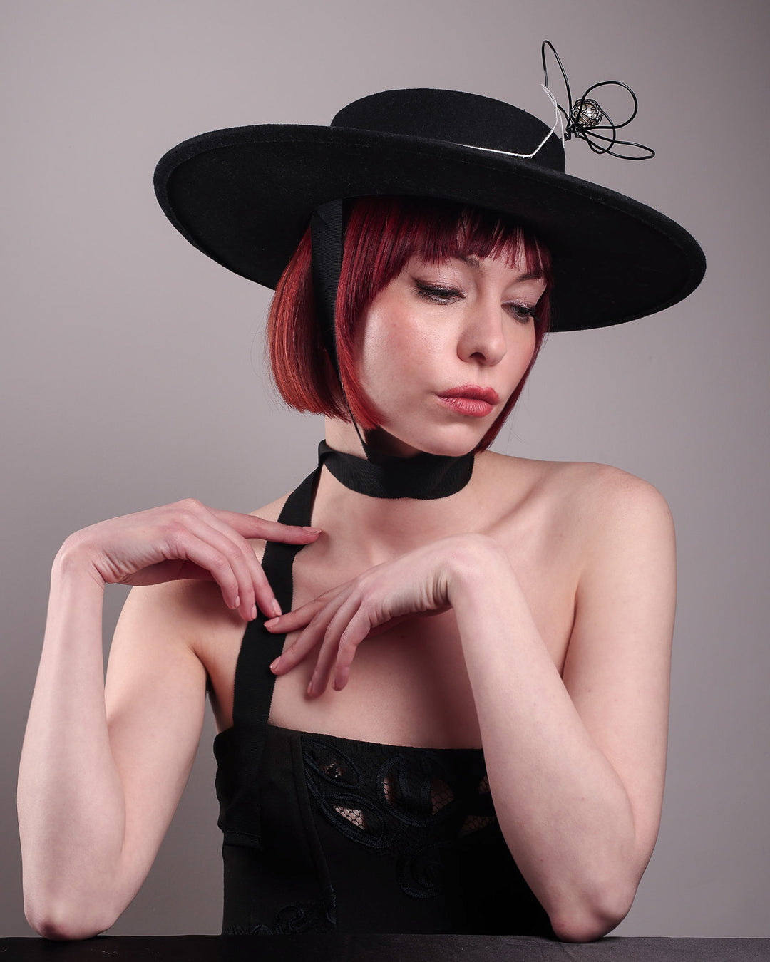 The model showcases the front right side of the Lune wide-brim boater hat from the below angle, crafted in deep black felt, the hat's crown is detailed with a hand-stitched outline of a hand—an abstract nod to surrealist elegance—encircling a wire ornament that loops skyward like an unruly ribbon flower or whispered thought. Designed by Merve Bayindir.