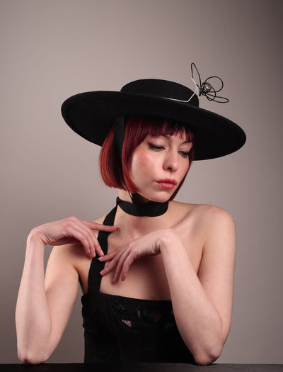 The model showcases the front right side of the Lune wide-brim boater hat from the below angle, crafted in deep black felt, the hat's crown is detailed with a hand-stitched outline of a hand—an abstract nod to surrealist elegance—encircling a wire ornament that loops skyward like an unruly ribbon flower or whispered thought. Designed by Merve Bayindir.