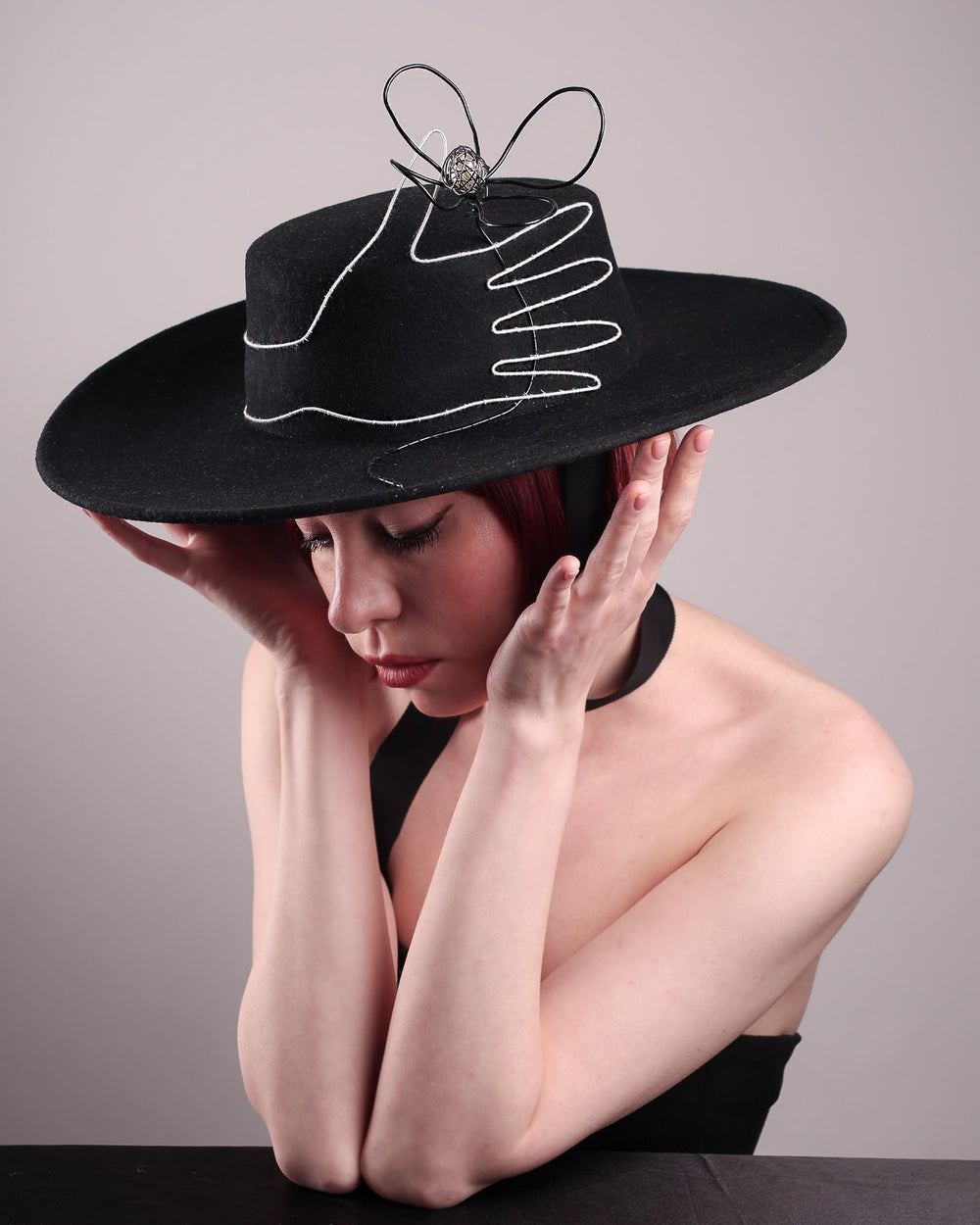 The model showcases the left  front side from the above angle of the Lune wide-brim boater hat, crafted in deep black felt, the hat's crown is detailed with a hand-stitched outline of a hand—an abstract nod to surrealist elegance—encircling a wire ornament that loops skyward like an unruly ribbon flower or whispered thought. Designed by Merve Bayindir.
