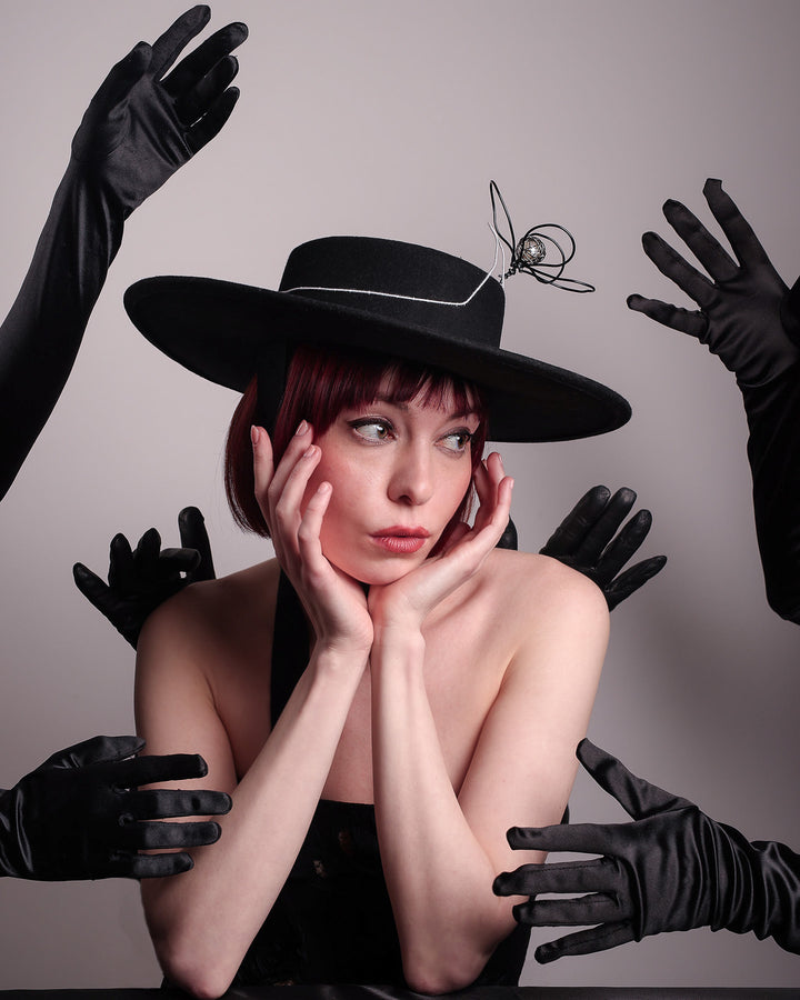 Editorial picture with a a lot of glowed hand surrounding of the model showcases the front of the Lune wide-brim boater hat, crafted in deep black felt, the hat&
