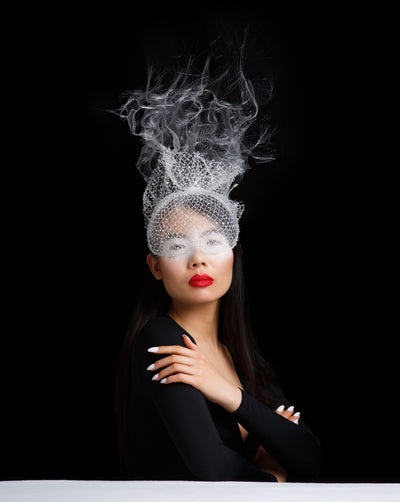 Model is wearing a mesh half face covering white mask covered with sequinned veil, back of the mask has goes up with teared crinoline creating a cloud like affect.