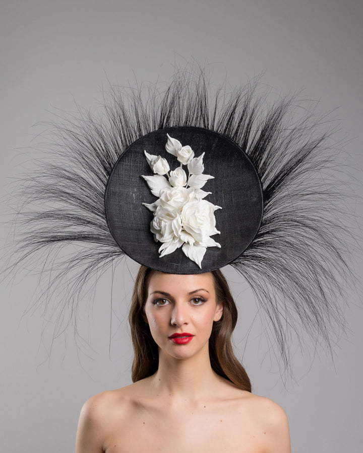 The model is showcasing the front side close up of the dramatic, sculptural fascinator featuring a sleek black sinamay disc base. From the edges, long, fine black quills radiate outward in a striking sunburst effect, creating movement and sharp elegance. At the center sits a cluster of handcrafted ivory roses and buds with matching leaves, providing a contrast against the dark base. Designed and made by Merve Bayindir Millinery.
