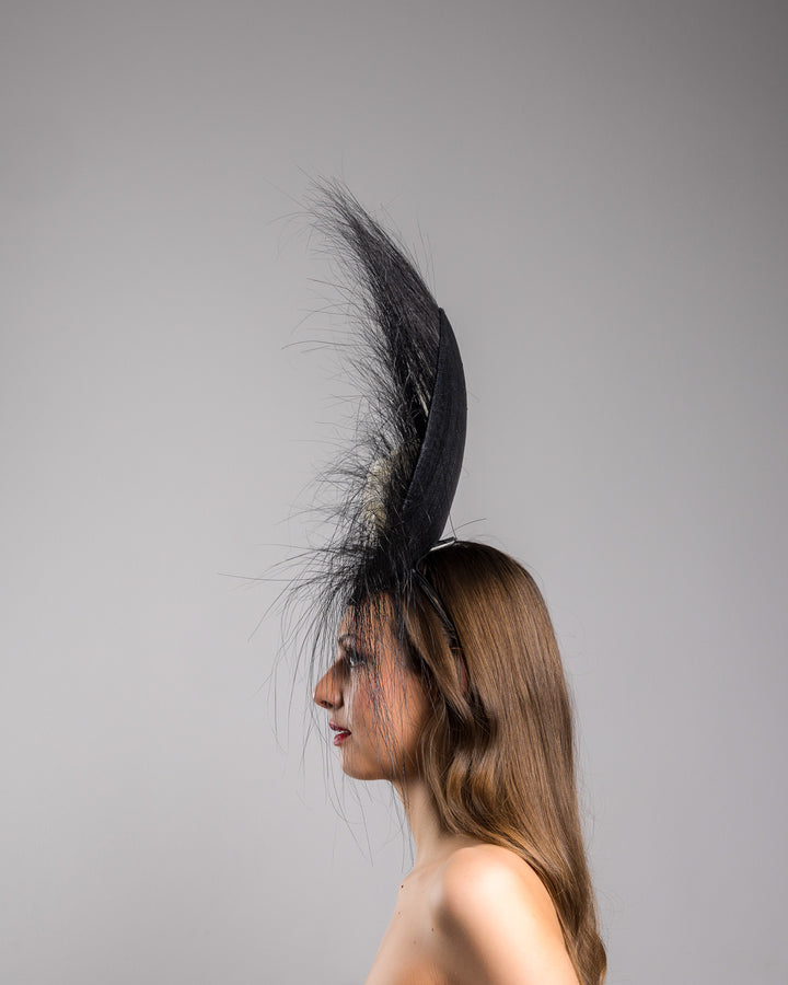 The model is showcasing the left side close up of the dramatic, sculptural fascinator featuring a sleek black sinamay disc base. From the edges, long, fine black quills radiate outward in a striking sunburst effect, creating movement and sharp elegance. At the center sits a cluster of handcrafted ivory roses and buds with matching leaves, providing a contrast against the dark base. Designed and made by Merve Bayindir Millinery.
