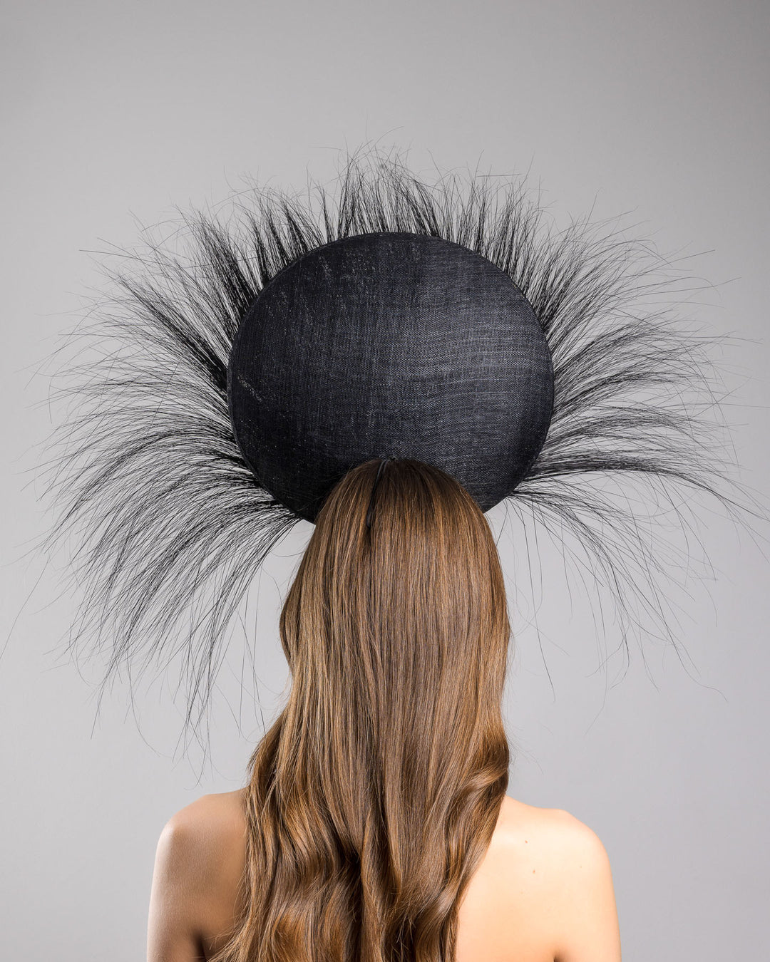 The model is showcasing the back side close up of the dramatic, sculptural fascinator featuring a sleek black sinamay disc base. From the edges, long, fine black quills radiate outward in a striking sunburst effect, creating movement and sharp elegance. At the center sits a cluster of handcrafted ivory roses and buds with matching leaves, providing a contrast against the dark base. Designed and made by Merve Bayindir Millinery.
