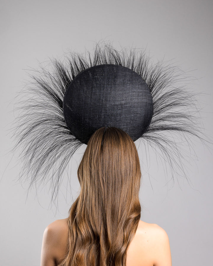The model is showcasing the back side close up of the dramatic, sculptural fascinator featuring a sleek black sinamay disc base. From the edges, long, fine black quills radiate outward in a striking sunburst effect, creating movement and sharp elegance. At the center sits a cluster of handcrafted ivory roses and buds with matching leaves, providing a contrast against the dark base. Designed and made by Merve Bayindir Millinery.
