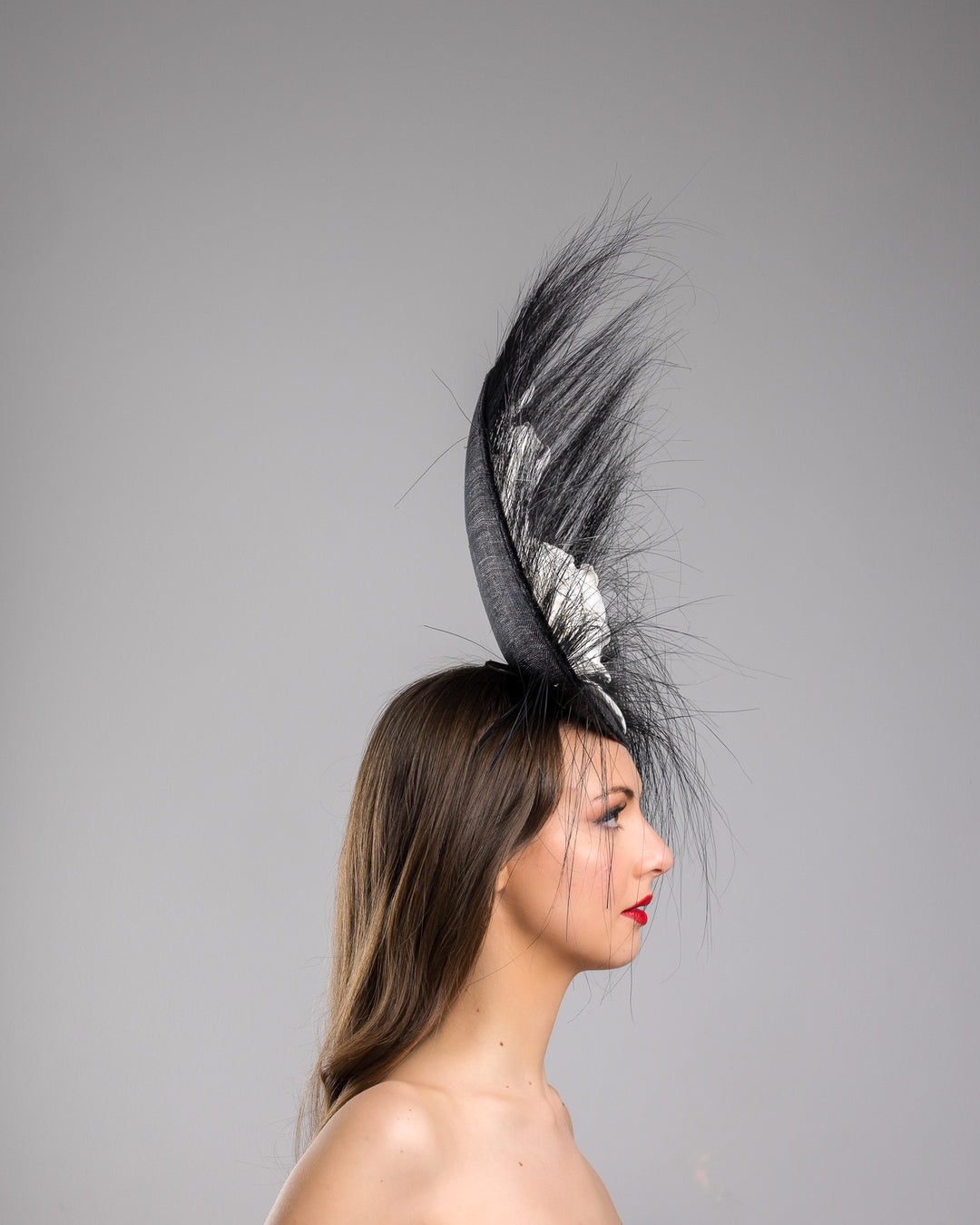 The model is showcasing the right side close up of the dramatic, sculptural fascinator featuring a sleek black sinamay disc base. From the edges, long, fine black quills radiate outward in a striking sunburst effect, creating movement and sharp elegance. At the center sits a cluster of handcrafted ivory roses and buds with matching leaves, providing a contrast against the dark base. Designed and made by Merve Bayindir Millinery.
