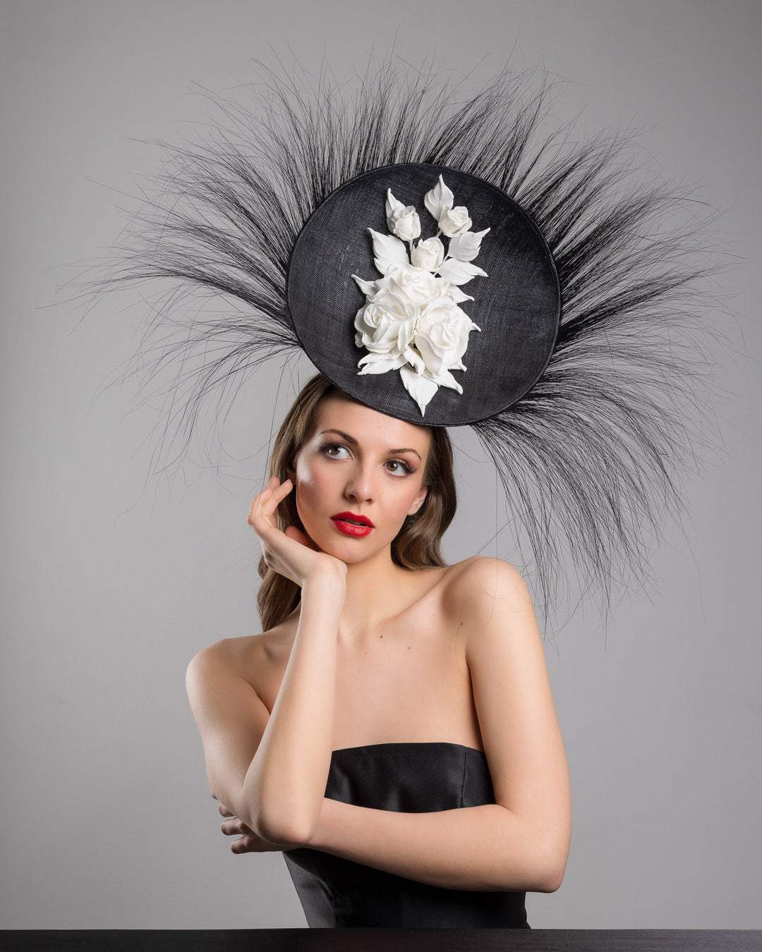 The female model is posing, wearing
Martinian, the dramatic, sculptural fascinator featuring a sleek black sinamay disc base. From the edges, long, fine black quills radiate outward in a striking sunburst effect, creating movement and sharp elegance. At the center sits a cluster of handcrafted ivory roses and buds with matching leaves, providing a contrast against the dark base. Designed and made by Merve Bayindir Millinery.
