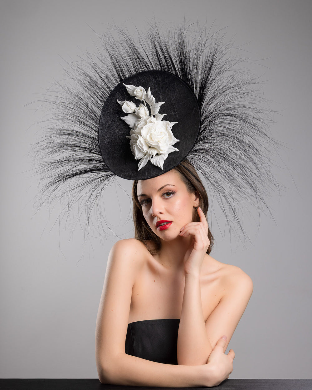 The female model is posing, wearing
Martinian, the dramatic, sculptural fascinator featuring a sleek black sinamay disc base. From the edges, long, fine black quills radiate outward in a striking sunburst effect, creating movement and sharp elegance. At the center sits a cluster of handcrafted ivory roses and buds with matching leaves, providing a contrast against the dark base. Designed and made by Merve Bayindir Millinery.
