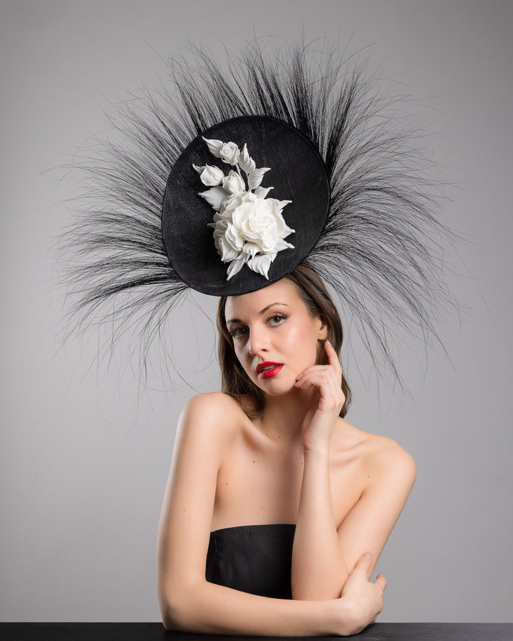 The female model is posing, wearing
Martinian, the dramatic, sculptural fascinator featuring a sleek black sinamay disc base. From the edges, long, fine black quills radiate outward in a striking sunburst effect, creating movement and sharp elegance. At the center sits a cluster of handcrafted ivory roses and buds with matching leaves, providing a contrast against the dark base. Designed and made by Merve Bayindir Millinery.
