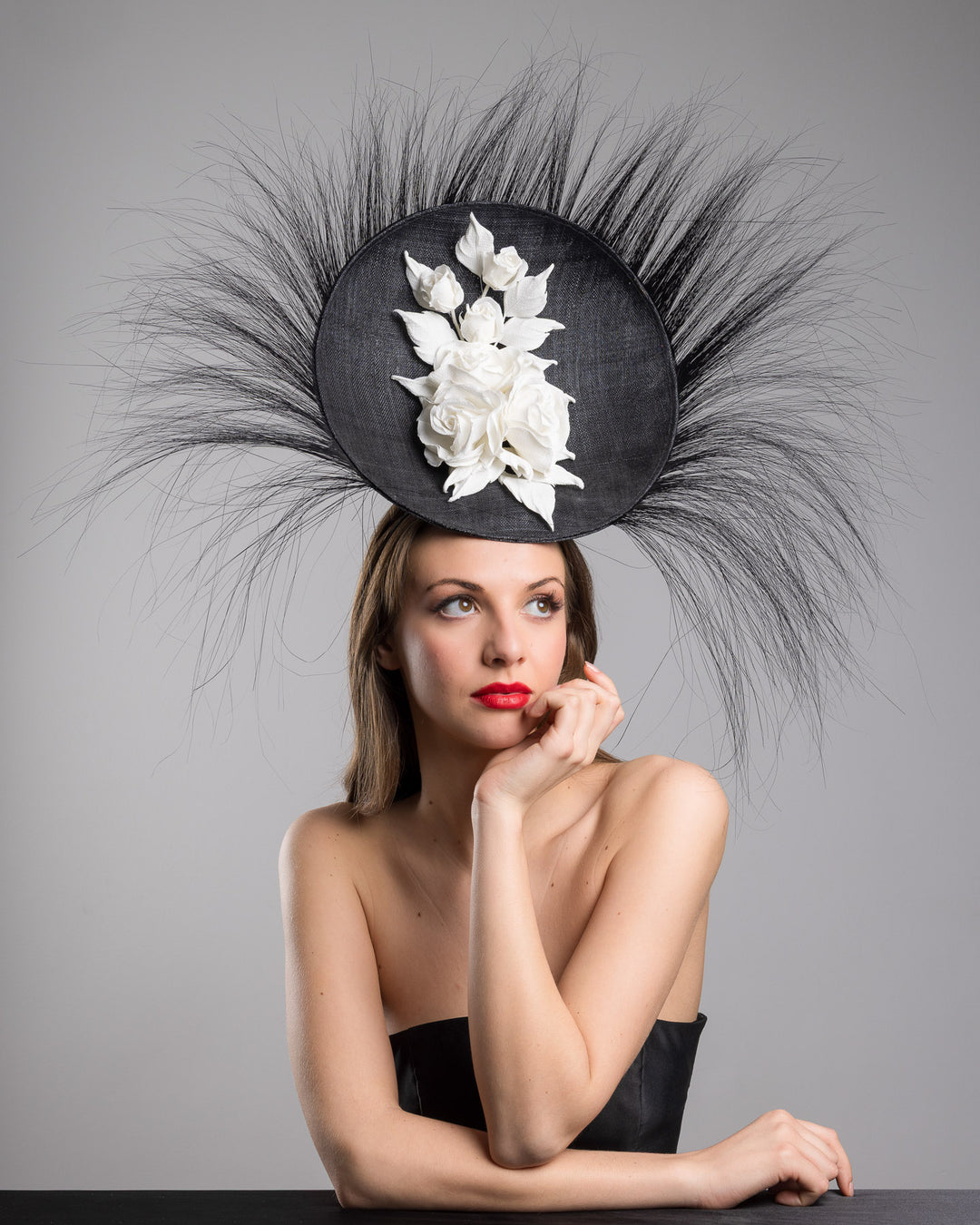 The female model is posing, wearing
Martinian, the dramatic, sculptural fascinator featuring a sleek black sinamay disc base. From the edges, long, fine black quills radiate outward in a striking sunburst effect, creating movement and sharp elegance. At the center sits a cluster of handcrafted ivory roses and buds with matching leaves, providing a contrast against the dark base. Designed and made by Merve Bayindir Millinery.

