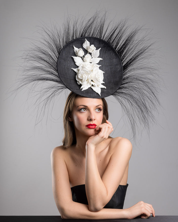 The female model is posing, wearing
Martinian, the dramatic, sculptural fascinator featuring a sleek black sinamay disc base. From the edges, long, fine black quills radiate outward in a striking sunburst effect, creating movement and sharp elegance. At the center sits a cluster of handcrafted ivory roses and buds with matching leaves, providing a contrast against the dark base. Designed and made by Merve Bayindir Millinery.
