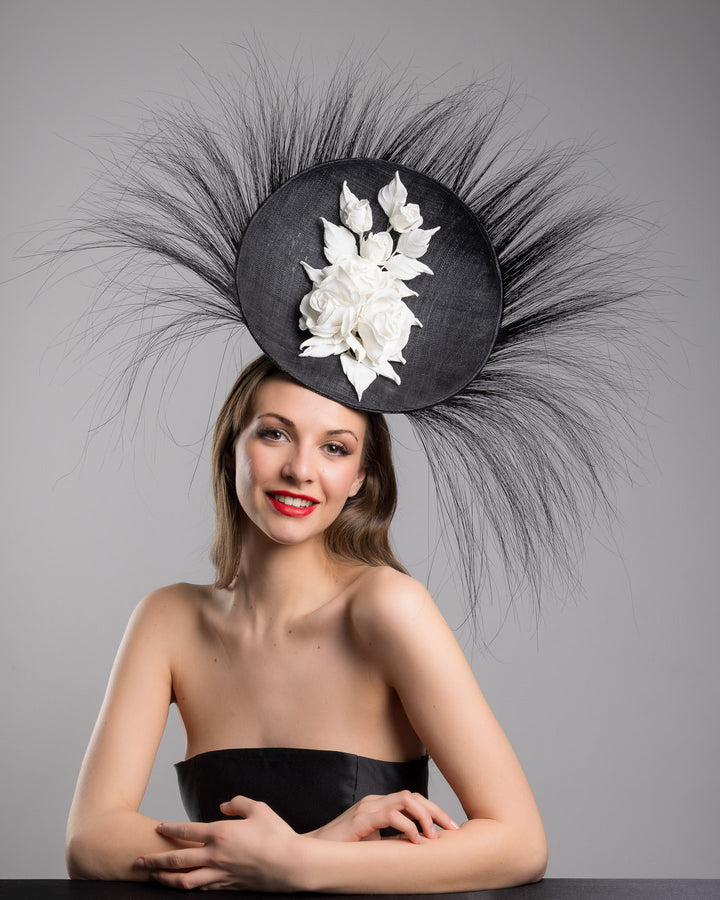 The female model is posing, wearing
Martinian, the dramatic, sculptural fascinator featuring a sleek black sinamay disc base. From the edges, long, fine black quills radiate outward in a striking sunburst effect, creating movement and sharp elegance. At the center sits a cluster of handcrafted ivory roses and buds with matching leaves, providing a contrast against the dark base. Designed and made by Merve Bayindir Millinery.
