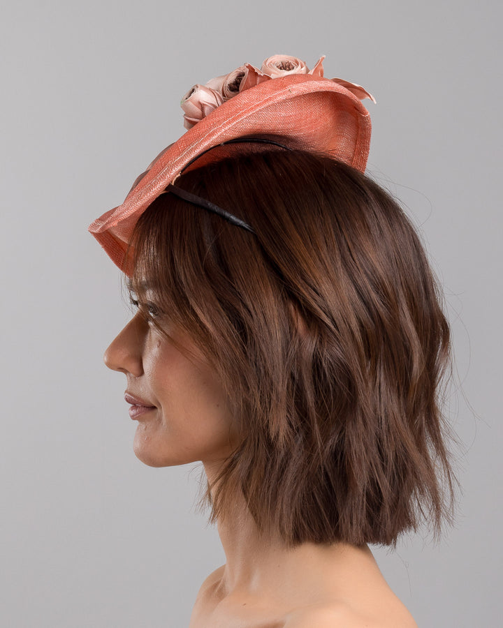The model is showcasing the left side close up of the coral sinamay fascinator with a softly sculpted asymmetrical base, topped with blush-toned handcrafted rosette flowers and delicate matching leaves, mounted on a slim headband. Designed and made by Merve Bayindir Millinery.
