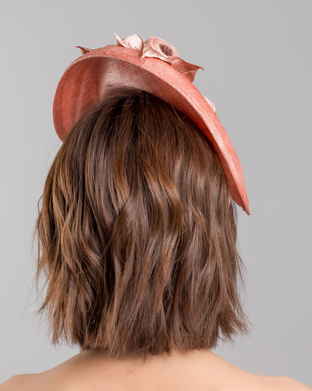 The model is showcasing the back side close up of the coral sinamay fascinator with a softly sculpted asymmetrical base, topped with blush-toned handcrafted rosette flowers and delicate matching leaves, mounted on a slim headband. Designed and made by Merve Bayindir Millinery.
