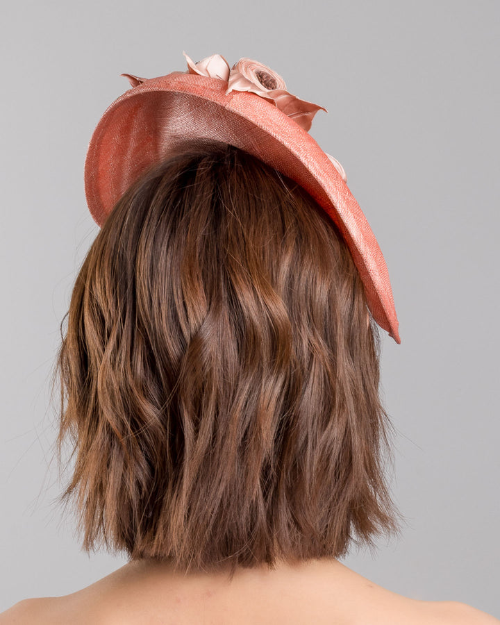 The model is showcasing the back side close up of the coral sinamay fascinator with a softly sculpted asymmetrical base, topped with blush-toned handcrafted rosette flowers and delicate matching leaves, mounted on a slim headband. Designed and made by Merve Bayindir Millinery.
