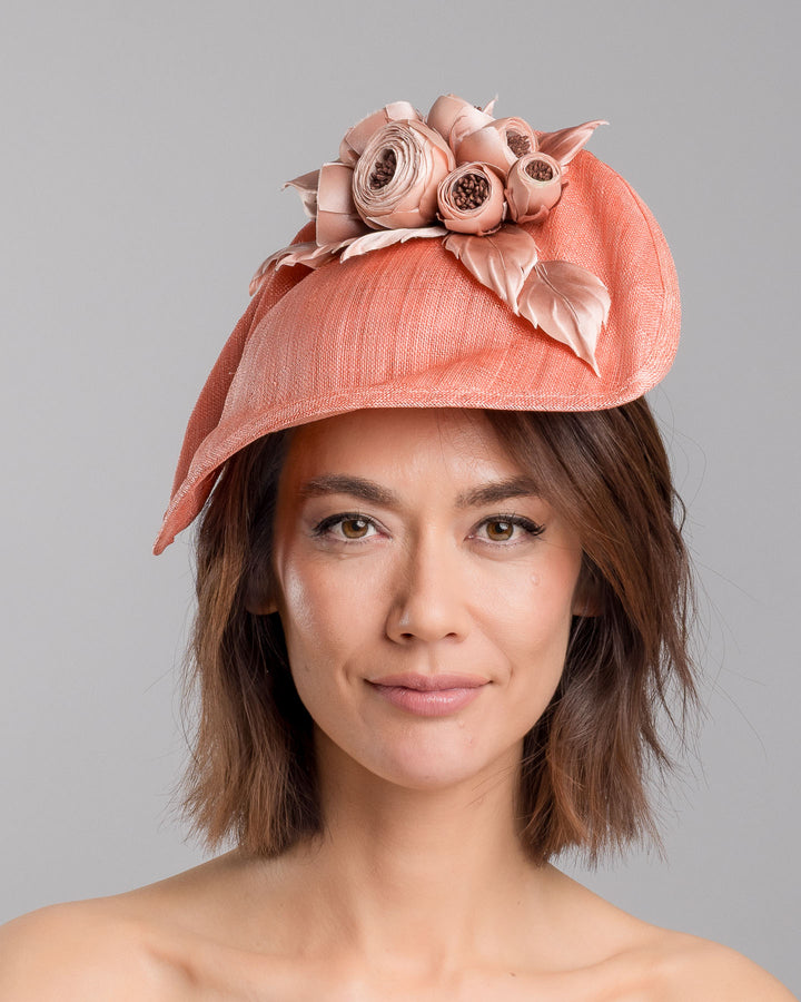 The model is showcasing the front side close up of the coral sinamay fascinator with a softly sculpted asymmetrical base, topped with blush-toned handcrafted rosette flowers and delicate matching leaves, mounted on a slim headband. Designed and made by Merve Bayindir Millinery.
