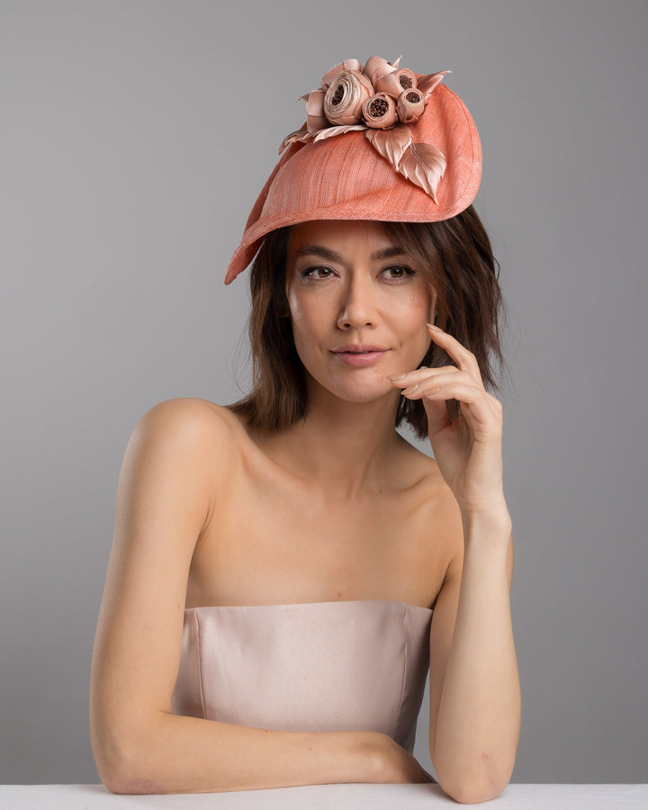 The female model is posing, wearing
Merrit, the coral sinamay fascinator with a softly sculpted asymmetrical base, topped with blush-toned handcrafted rosette flowers and delicate matching leaves, mounted on a slim headband. Designed and made by Merve Bayindir Millinery.
