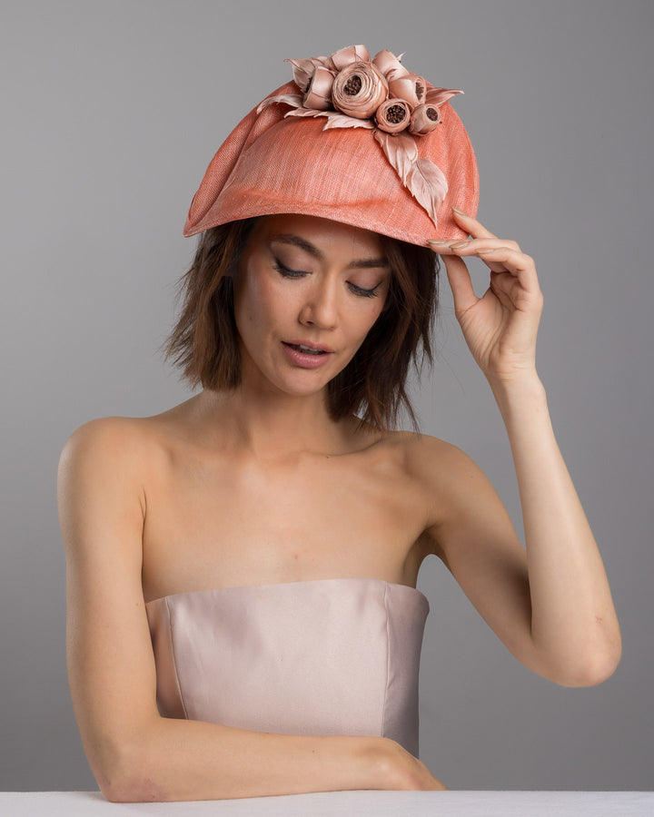 The female model is posing, wearing
Merrit, the coral sinamay fascinator with a softly sculpted asymmetrical base, topped with blush-toned handcrafted rosette flowers and delicate matching leaves, mounted on a slim headband. Designed and made by Merve Bayindir Millinery.
