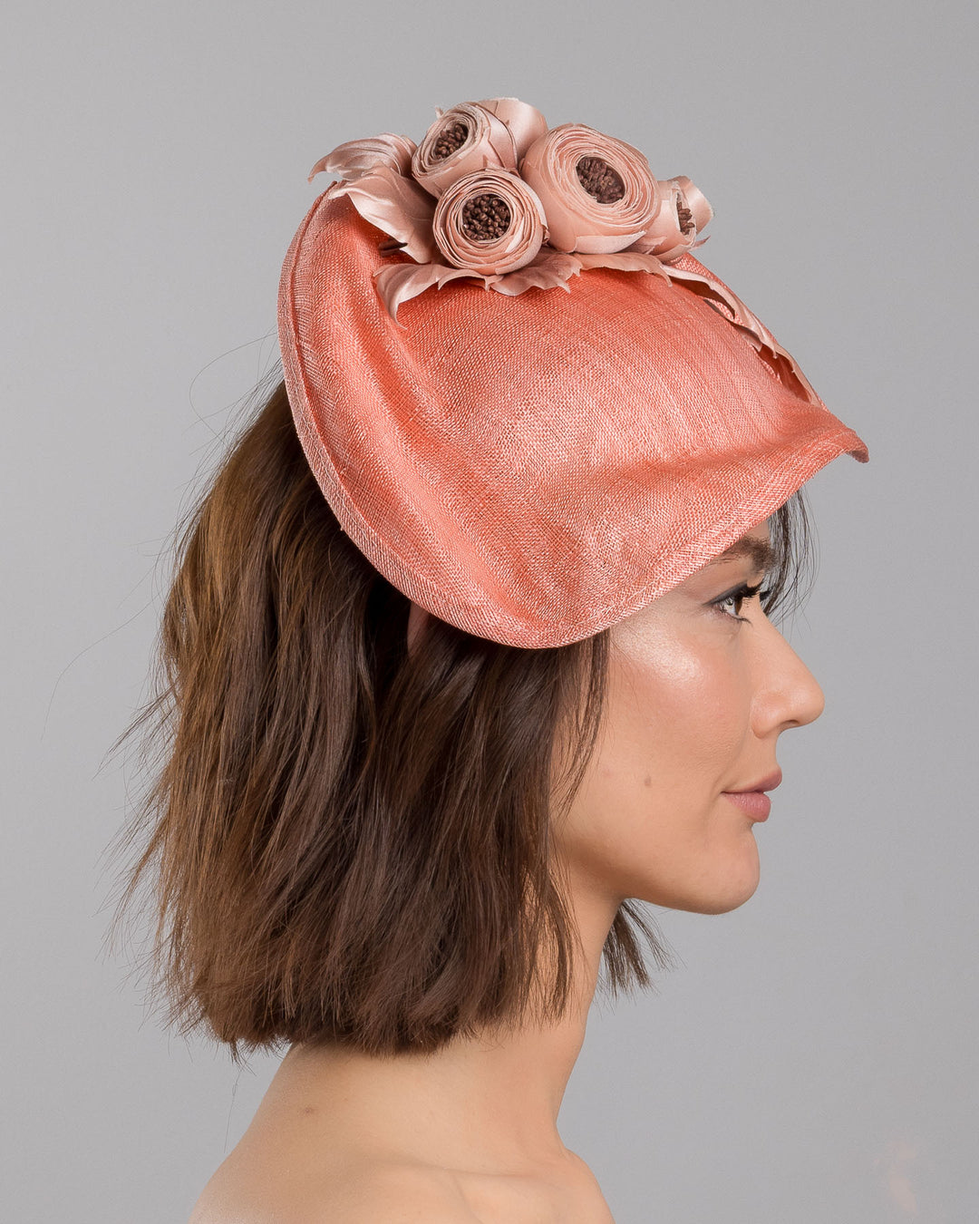 The model is showcasing the right side close up of the coral sinamay fascinator with a softly sculpted asymmetrical base, topped with blush-toned handcrafted rosette flowers and delicate matching leaves, mounted on a slim headband. Designed and made by Merve Bayindir Millinery.

