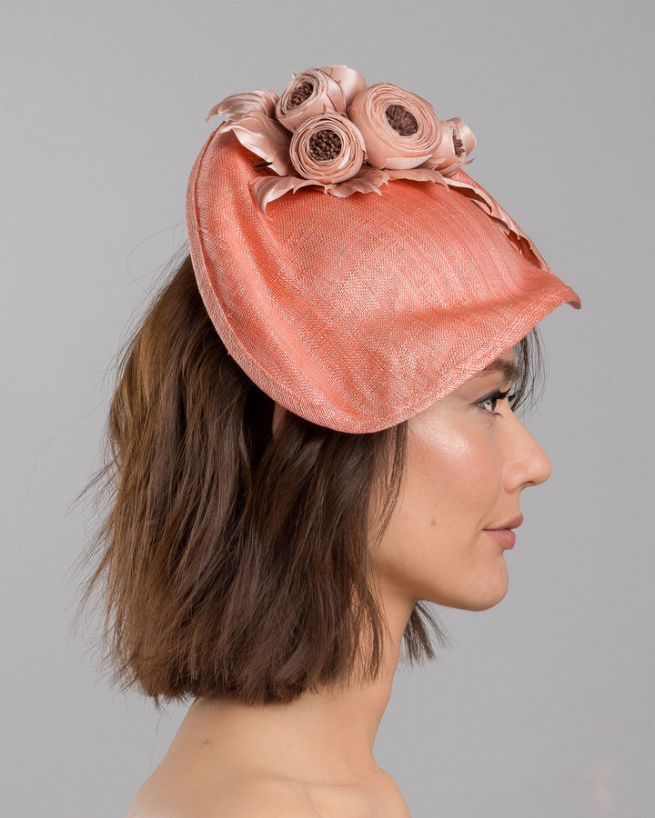 The model is showcasing the right side close up of the coral sinamay fascinator with a softly sculpted asymmetrical base, topped with blush-toned handcrafted rosette flowers and delicate matching leaves, mounted on a slim headband. Designed and made by Merve Bayindir Millinery.
