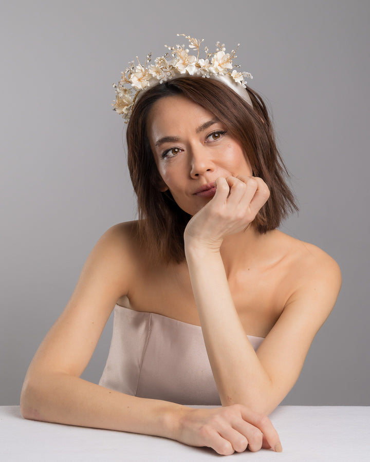 The female model is posing, wearing Mirel, the ivory satin padded headband featuring a crown-like arrangement of handcrafted translucent ivory flowers. The design is accented with fine gold branches, crystal details, pearl-like beads, and subtle grey leaf elements, creating a luminous and romantic statement piece ideal for brides and elegant formal occasions. Designed and made by Merve Bayindir Millinery.

