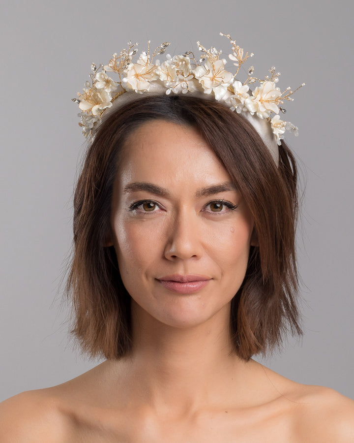 The model is showcasing the front side close up of the ivory satin padded headband featuring a crown-like arrangement of handcrafted translucent ivory flowers. The design is accented with fine gold branches, crystal details, pearl-like beads, and subtle grey leaf elements, creating a luminous and romantic statement piece ideal for brides and elegant formal occasions. Designed and made by Merve Bayindir Millinery.
