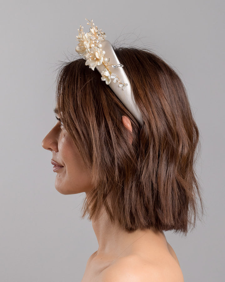 The model is showcasing the left side close up of the ivory satin padded headband featuring a crown-like arrangement of handcrafted translucent ivory flowers. The design is accented with fine gold branches, crystal details, pearl-like beads, and subtle grey leaf elements, creating a luminous and romantic statement piece ideal for brides and elegant formal occasions. Designed and made by Merve Bayindir Millinery.
