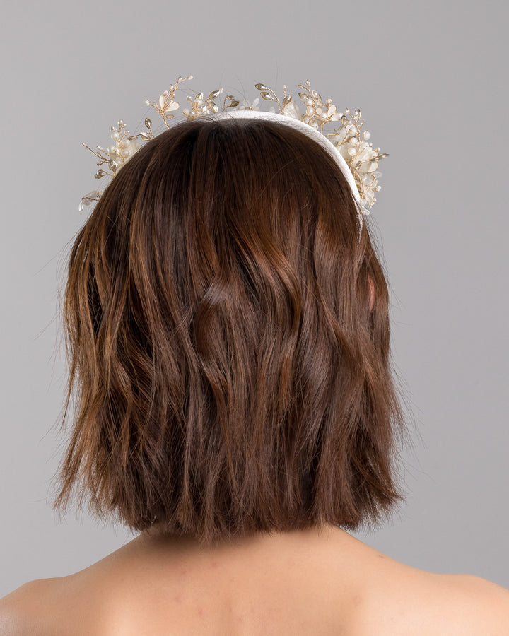 The model is showcasing the back side close up of the ivory satin padded headband featuring a crown-like arrangement of handcrafted translucent ivory flowers. The design is accented with fine gold branches, crystal details, pearl-like beads, and subtle grey leaf elements, creating a luminous and romantic statement piece ideal for brides and elegant formal occasions. Designed and made by Merve Bayindir Millinery.
