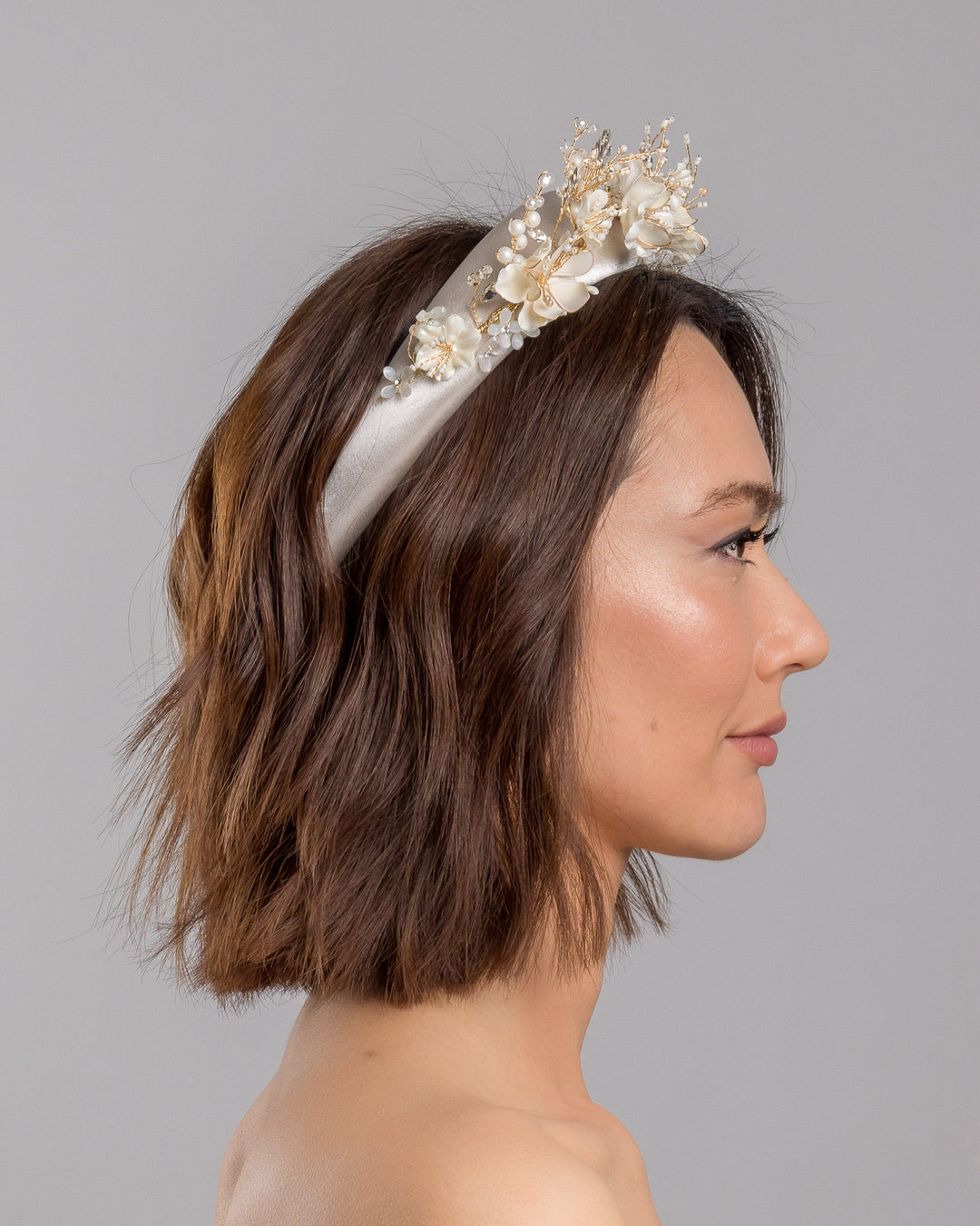The model is showcasing the right side close up of the ivory satin padded headband featuring a crown-like arrangement of handcrafted translucent ivory flowers. The design is accented with fine gold branches, crystal details, pearl-like beads, and subtle grey leaf elements, creating a luminous and romantic statement piece ideal for brides and elegant formal occasions. Designed and made by Merve Bayindir Millinery.
