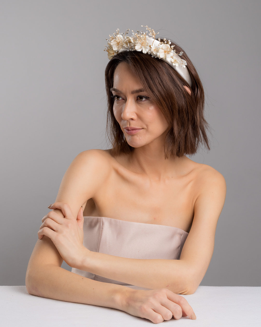 The female model is posing, wearing Mirel, the ivory satin padded headband featuring a crown-like arrangement of handcrafted translucent ivory flowers. The design is accented with fine gold branches, crystal details, pearl-like beads, and subtle grey leaf elements, creating a luminous and romantic statement piece ideal for brides and elegant formal occasions. Designed and made by Merve Bayindir Millinery.
