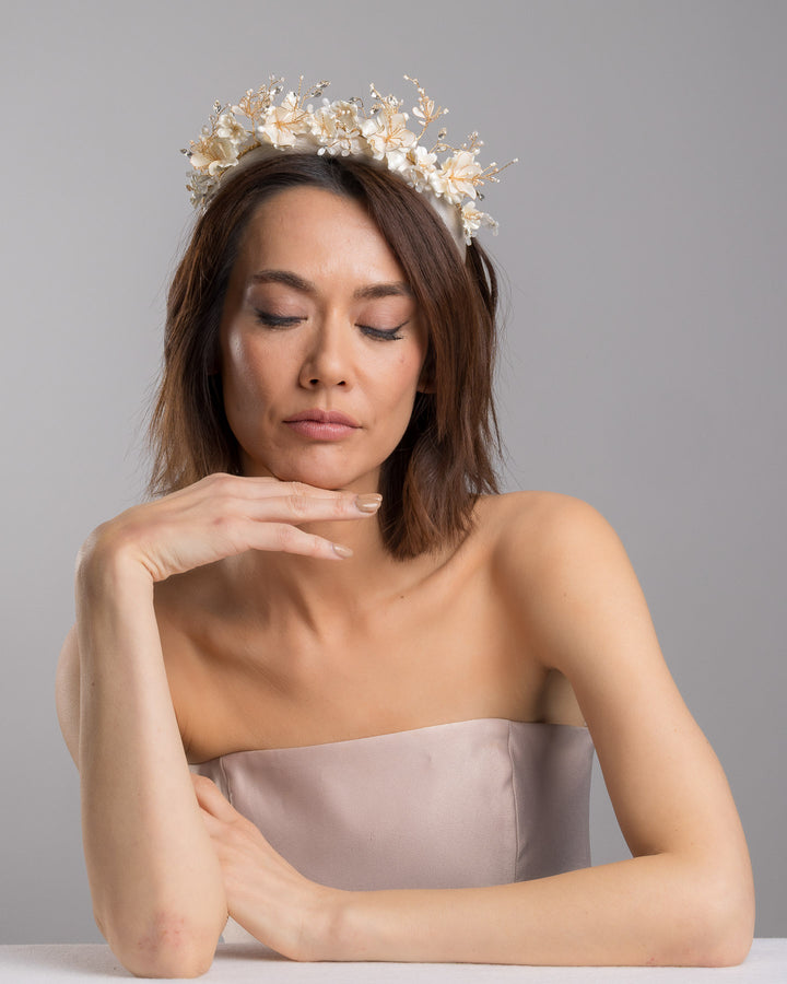 The female model is posing, wearing Mirel, the ivory satin padded headband featuring a crown-like arrangement of handcrafted translucent ivory flowers. The design is accented with fine gold branches, crystal details, pearl-like beads, and subtle grey leaf elements, creating a luminous and romantic statement piece ideal for brides and elegant formal occasions. Designed and made by Merve Bayindir Millinery.
