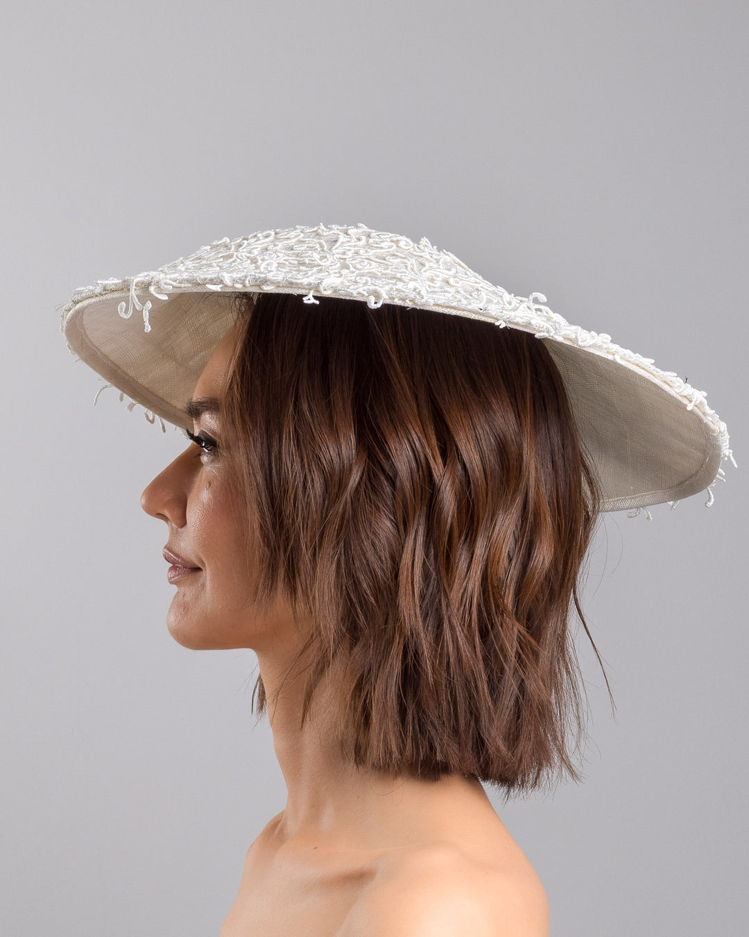 Model is showcasing the left side close up of white coloured wide brim white hat embellished with lace swirls. Designed and Made by Merve Bayindir Millinery.