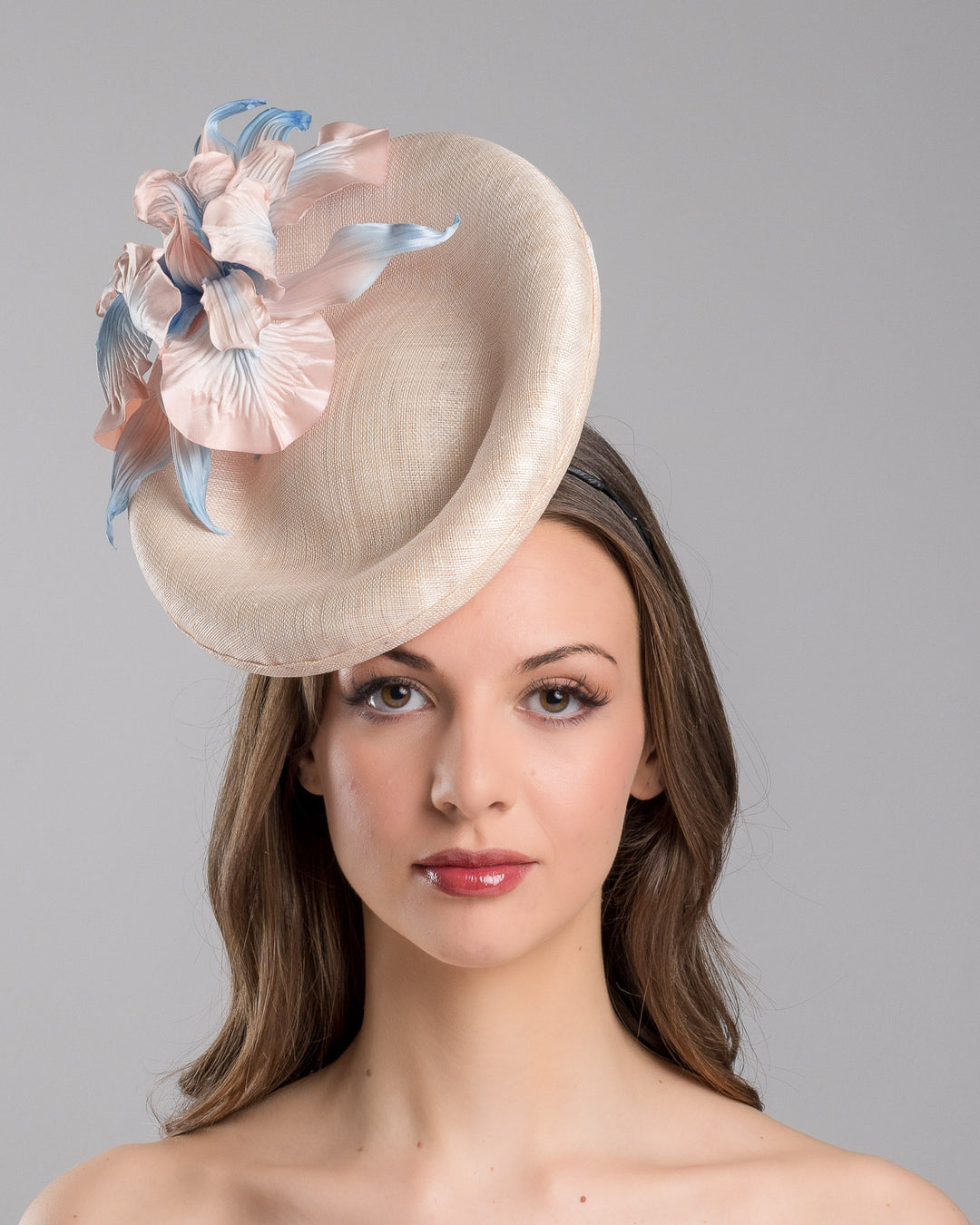 The model is showcasing the front side close up of the light pink oval high based fascinator decorated with custom made pink and blue shaded flowers. Designed and made by Merve Bayindir Millinery.
