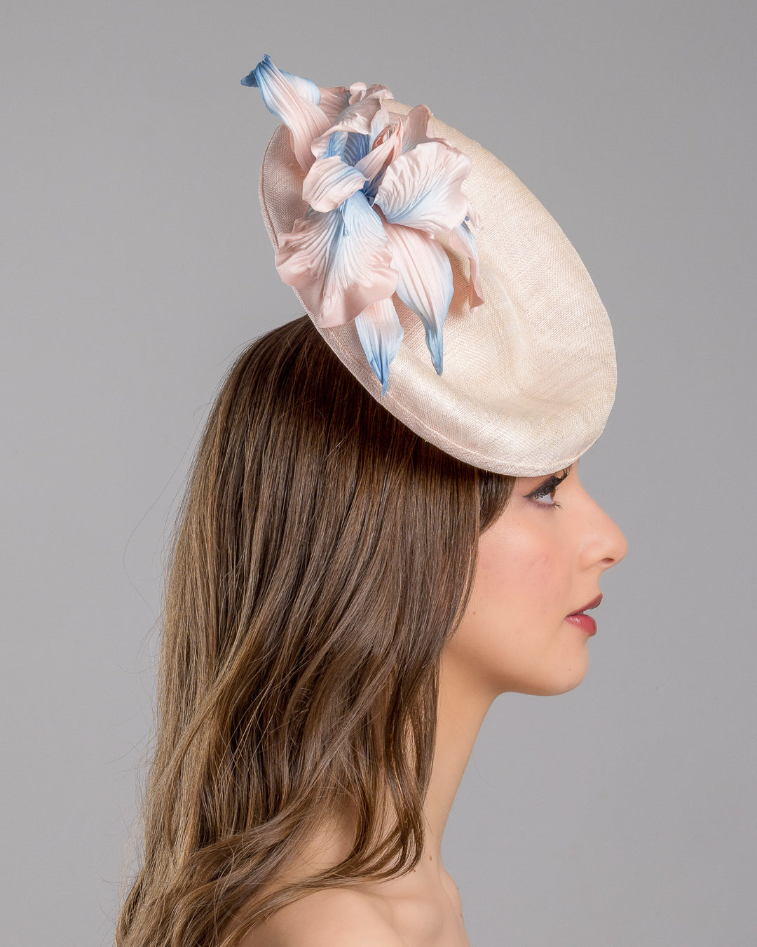 The model is showcasing the right side close up of the light pink oval high based fascinator decorated with custom made pink and blue shaded flowers. Designed and made by Merve Bayindir Millinery.
