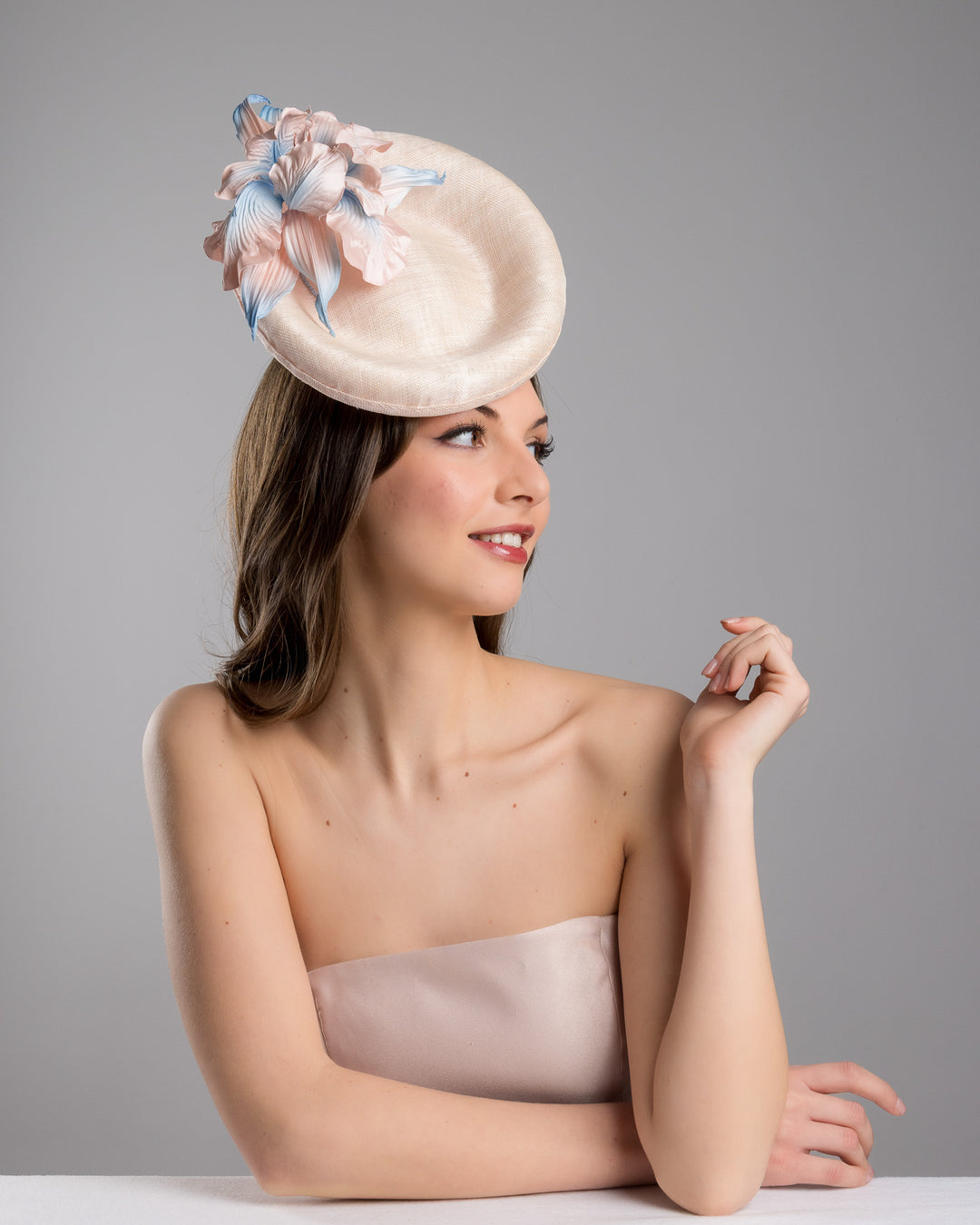 The female model is posing, wearing Marion,the light pink oval high based fascinator decorated with custom made pink and blue shaded flowers. Designed and made by Merve Bayindir Millinery.
